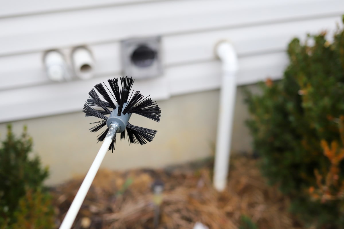 dryer vent cleaning brush with long handle with dryer vent opening on house in background