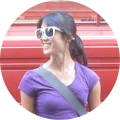Young brunette woman wearing sunglasses and a purple shirt with crossover strap smiling and looking toward left